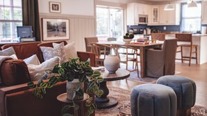 Un salon moderne avec un canapé, des poufs et une table basse. On y trouve également un coin repas avec une table et des chaises, et une cuisine à l’arrière-plan.