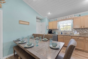 Un coin repas avec une table en bois dressée pour six personnes, une cuisine avec des placards en bois et une pancarte indiquant « MAISON DE PLAGE ».