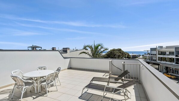 A rooftop terrace with a table and chairs, a view of the sea, and a building with an 'azura' sign.