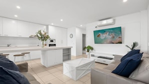 A modern kitchen with white cabinets, a central island, and a dining area with stools. There is a living room with a sofa, a coffee table, and a painting on the wall. - Paradiso 3 Bedroom Family 230 (Kingscliff)