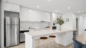A modern kitchen with white cabinetry, stainless steel appliances, and a central island with stools. - Paradiso 3 Bedroom Family 230 (Kingscliff)