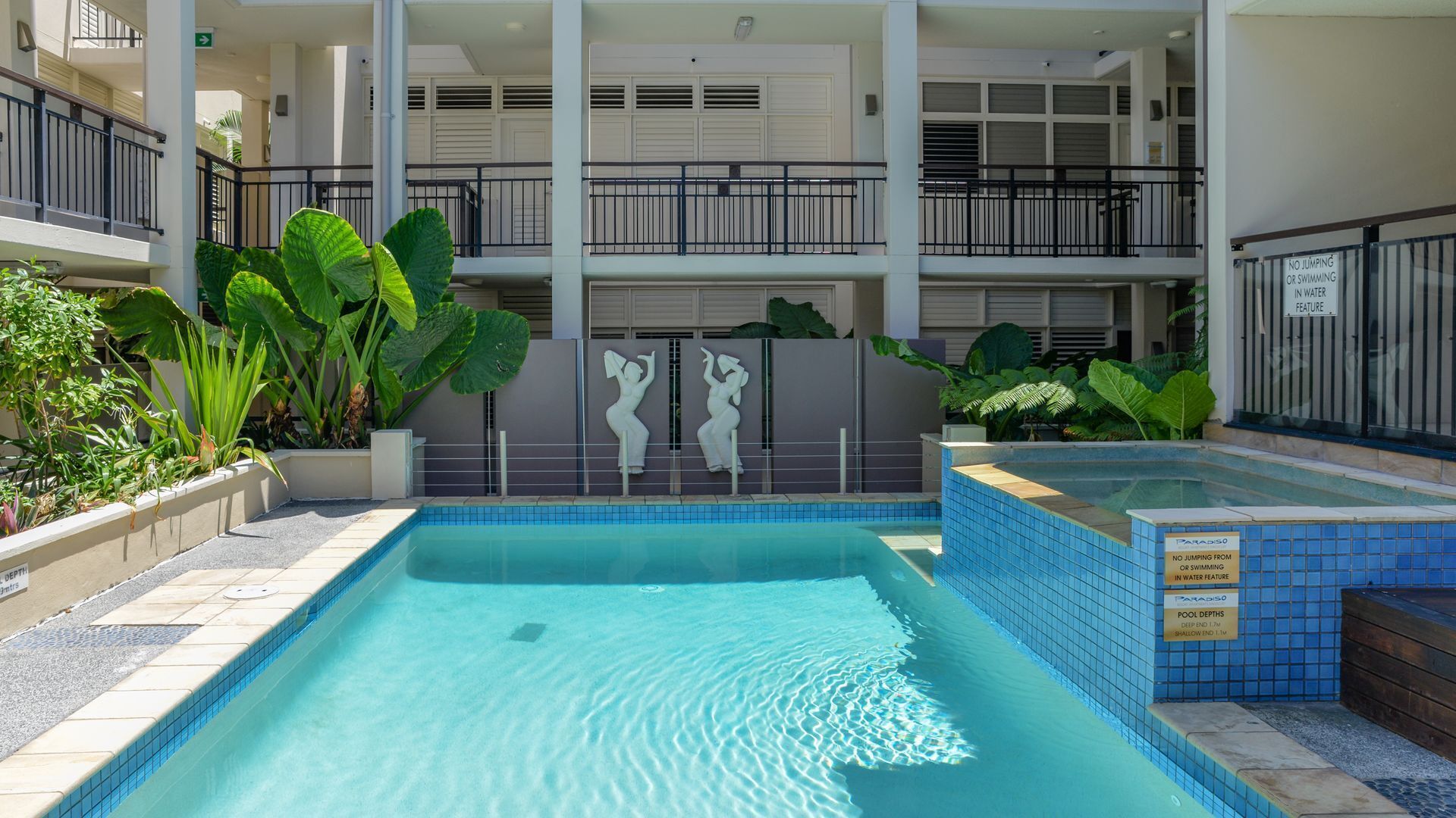 A swimming pool with a tiled edge, surrounded by plants and a building with balconies.