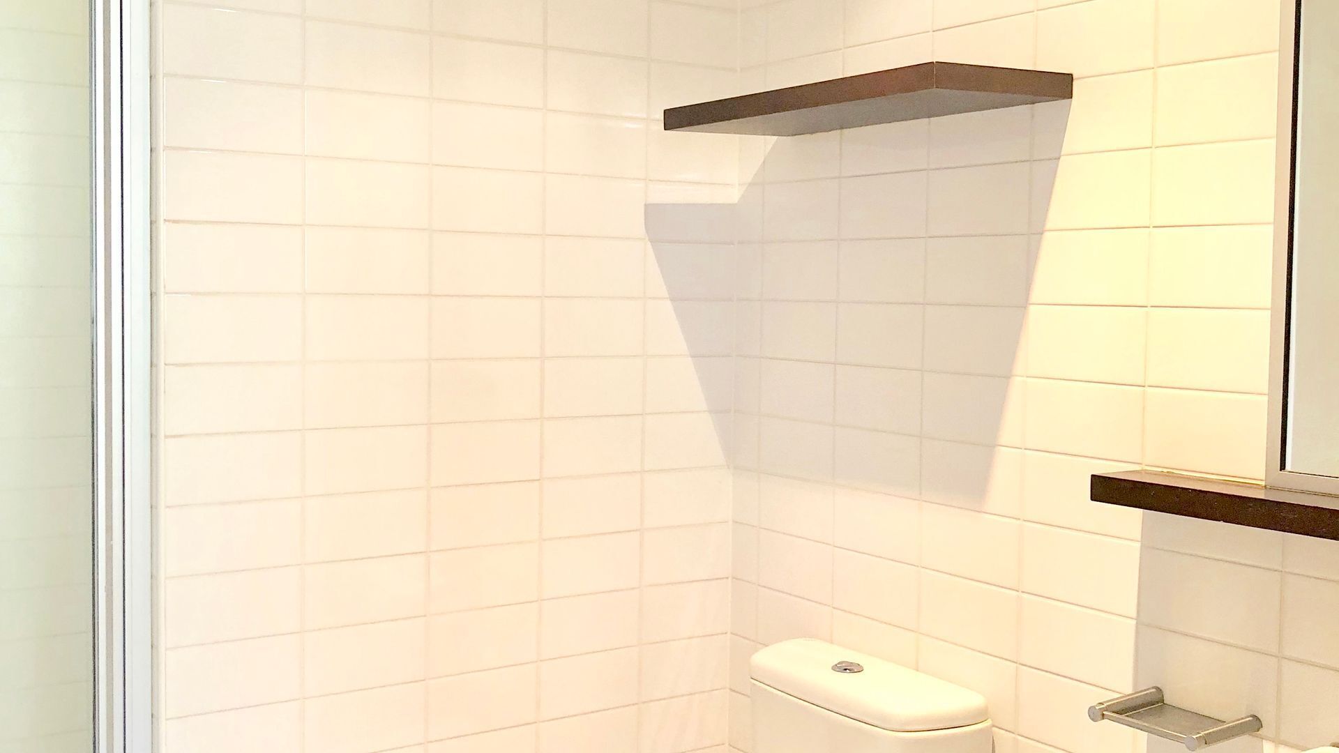 A bathroom with white tiled walls, a toilet, and a shower area with a shelf above the toilet.