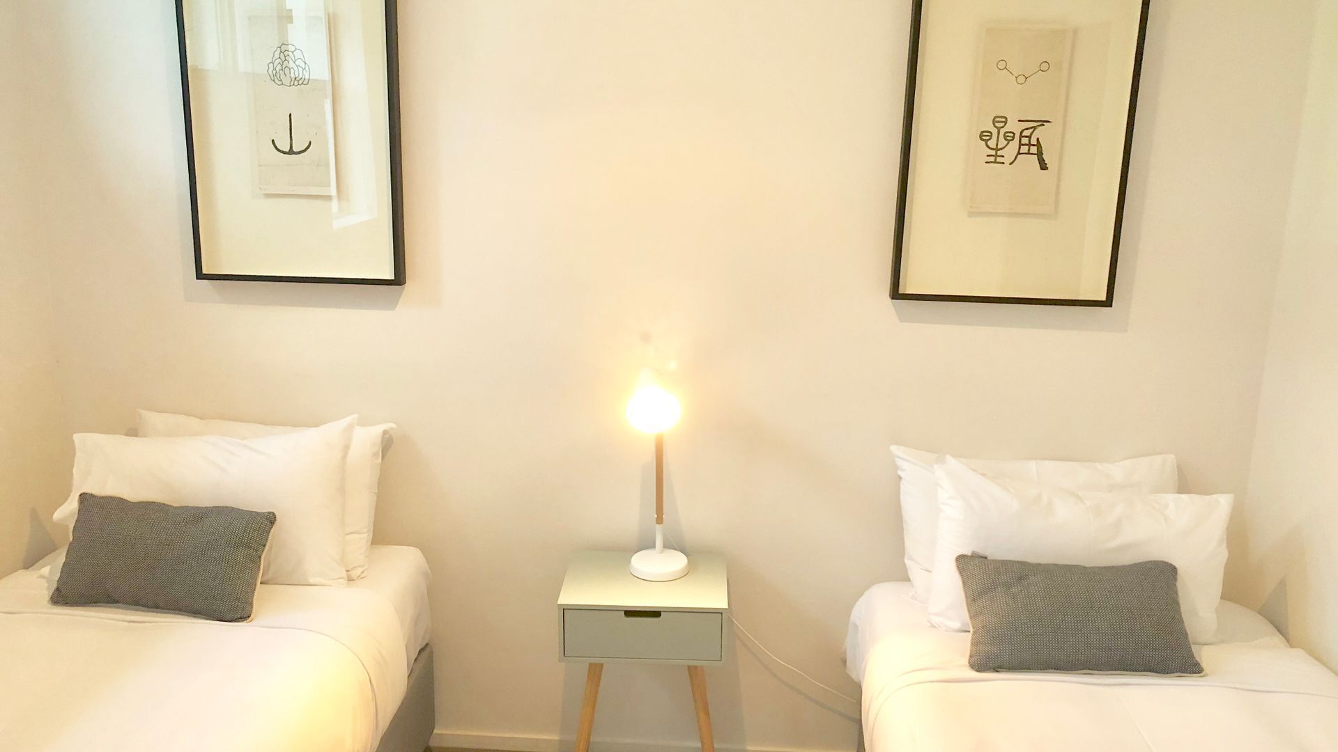 Une chambre d’hôtel avec deux lits, chacun agrémenté de draps blancs et d’un oreiller gris. Une petite table de chevet avec une lampe se trouve entre les deux lits. Les murs sont de couleur unie, et deux tableaux encadrés sont accrochés au-dessus des lits.