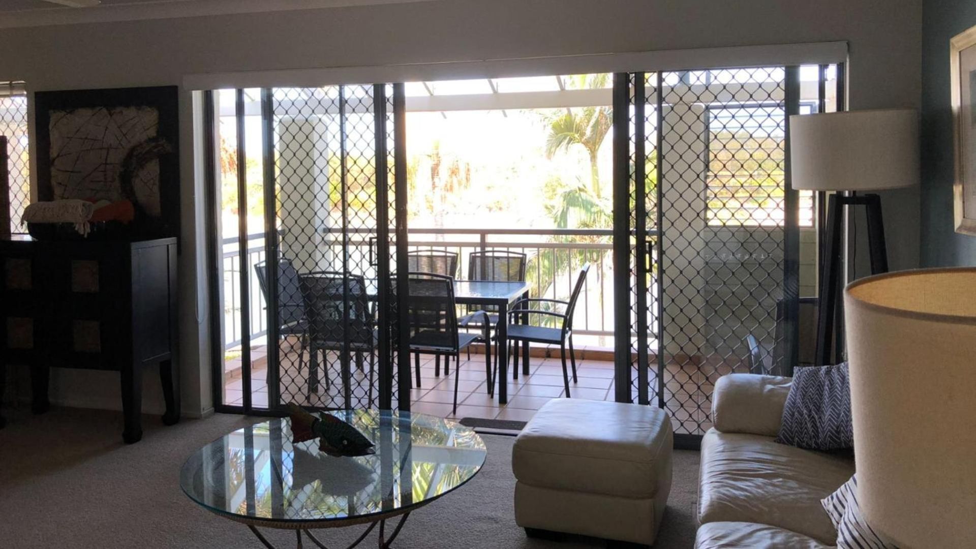 Un salon avec une table basse en verre, un canapé, une lampe et une vue sur une terrasse extérieure, accessible par une porte moustiquaire, où se trouvent une table et des chaises.