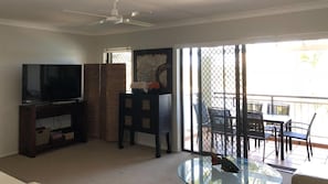 A living room with a television, a dark wooden cabinet, a glass coffee table, and a balcony with outdoor furniture.