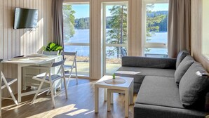 Une pièce chaleureuse avec un plancher en bois, un canapé gris, une petite table avec une plante, un téléviseur fixé au mur et de grandes fenêtres donnant sur un lac et des arbres.