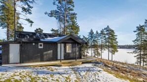 Une petite cabane en bois sombre, avec une seule porte et une seule fenêtre, située sur une pente enneigée, d’où l’on peut admirer un lac gelé et les arbres environnants.