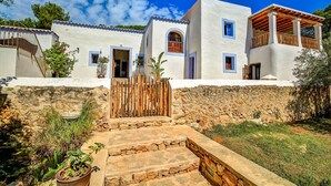 A traditional white building with blue accents, stone walls, and a wooden staircase leading to the entrance. - Awesome home in Sant Antoni de Portman (Sant Antoni de Portman)
