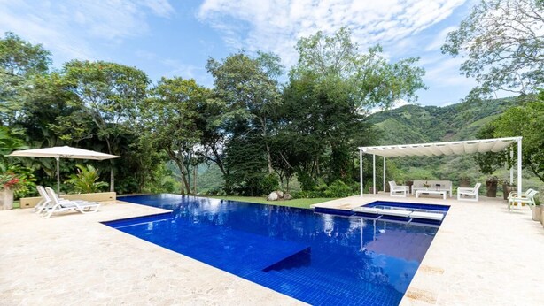 Una piscina rectangular con superficie azul cubierta de azulejos, rodeada por una terraza de piedra. Hay tumbonas y una zona de estar cubierta con un toldo blanco.