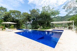 Ein rechteckiger Swimmingpool mit blauer Fliesenverkleidung, umgeben von einer Steindeckfläche. Liegestühle und eine überdachte Sitzgruppe mit weißem Baldachin sind vorhanden.