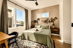 A bedroom with a bed, a desk, and a chair. There is a window with curtains, a wall-mounted mirror, and a wall with a horse mural.
