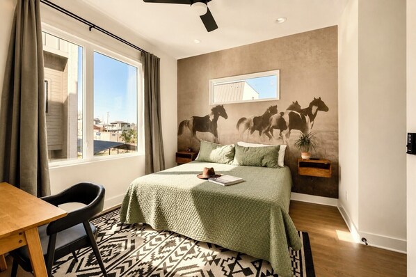 A bedroom with a bed, a desk, and a chair. There is a window with curtains, a wall-mounted mirror, and a wall with a horse mural.