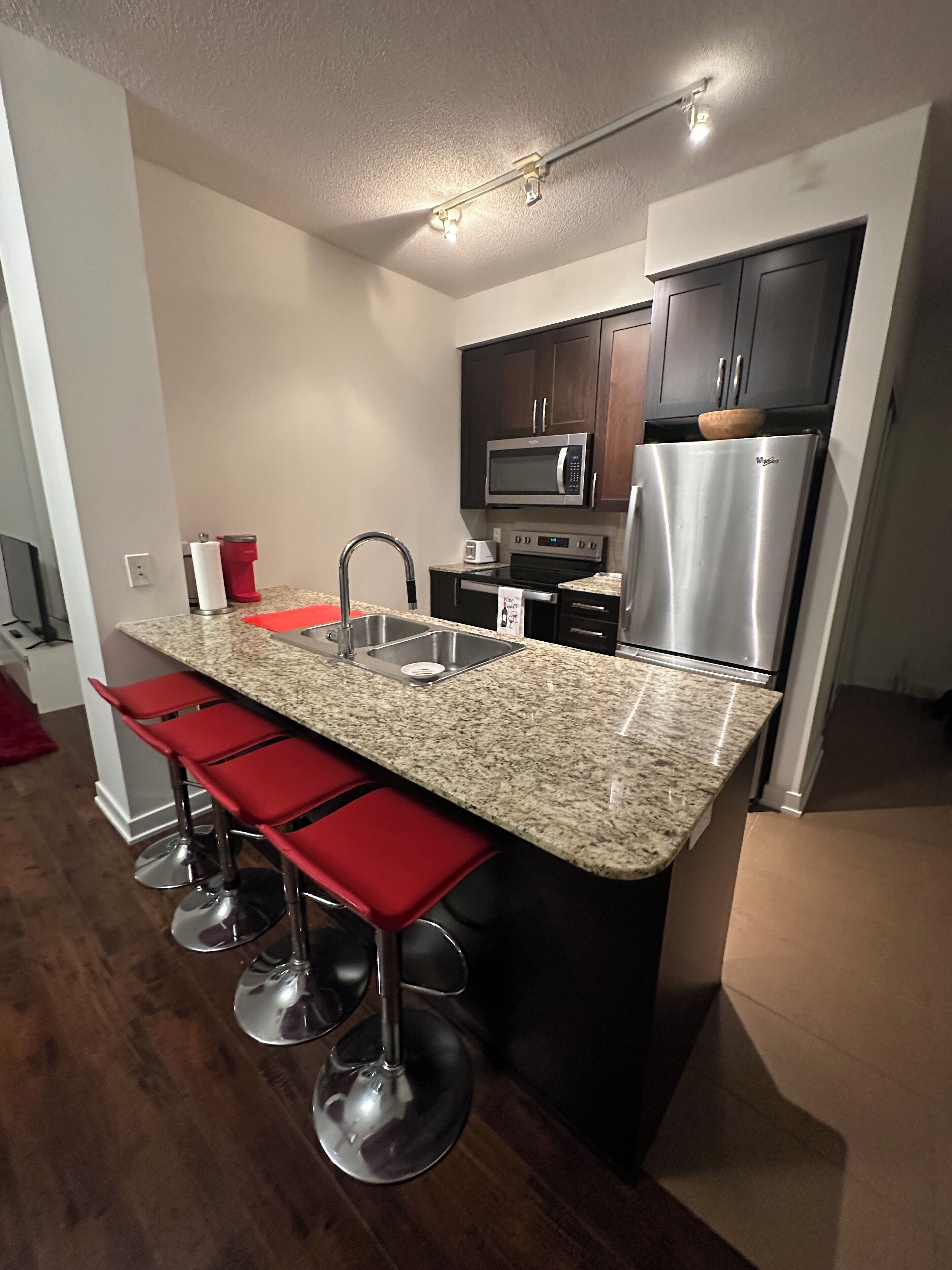 A modern kitchen with a granite countertop, stainless steel appliances, and red bar stools.