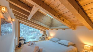 A bedroom with a large bed, wooden ceiling beams, a window with a view of trees, and a nightstand with a lamp. - Belvédère du Brévent - Welkeys (Chamonix-Mont-Blanc)