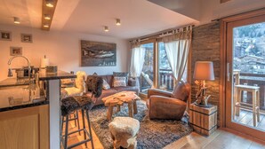 A cozy living area with a kitchen, a sofa, a coffee table, and a view of a snowy landscape through large windows. - Belvédère du Brévent - Welkeys (Chamonix-Mont-Blanc)