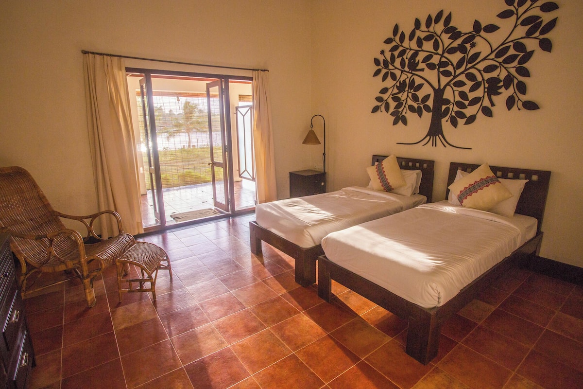A hotel room with two beds, a chair, a lamp, and a tree mural on the wall.