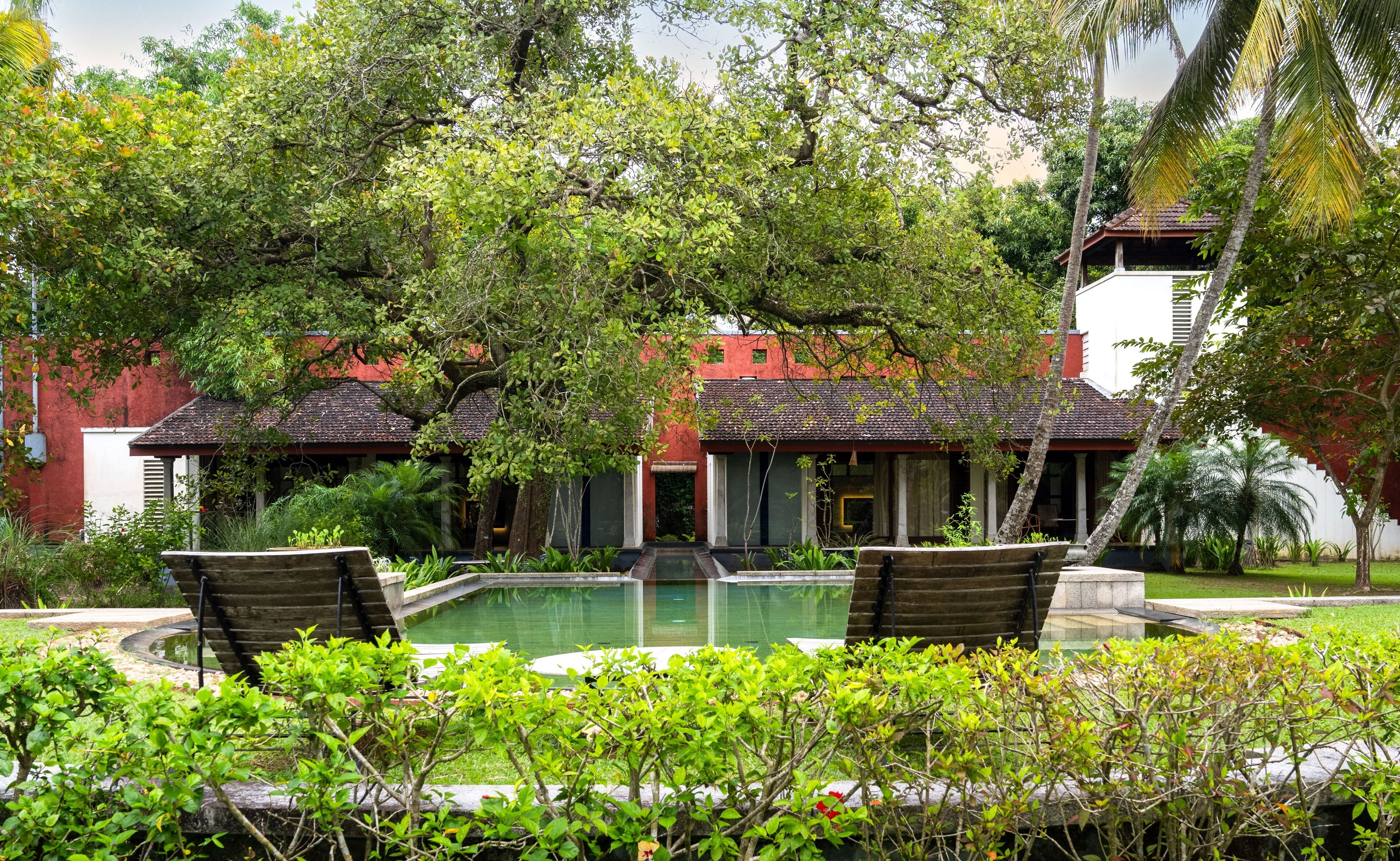 A pool area with lounge chairs, surrounded by lush greenery and a building with a tiled roof.