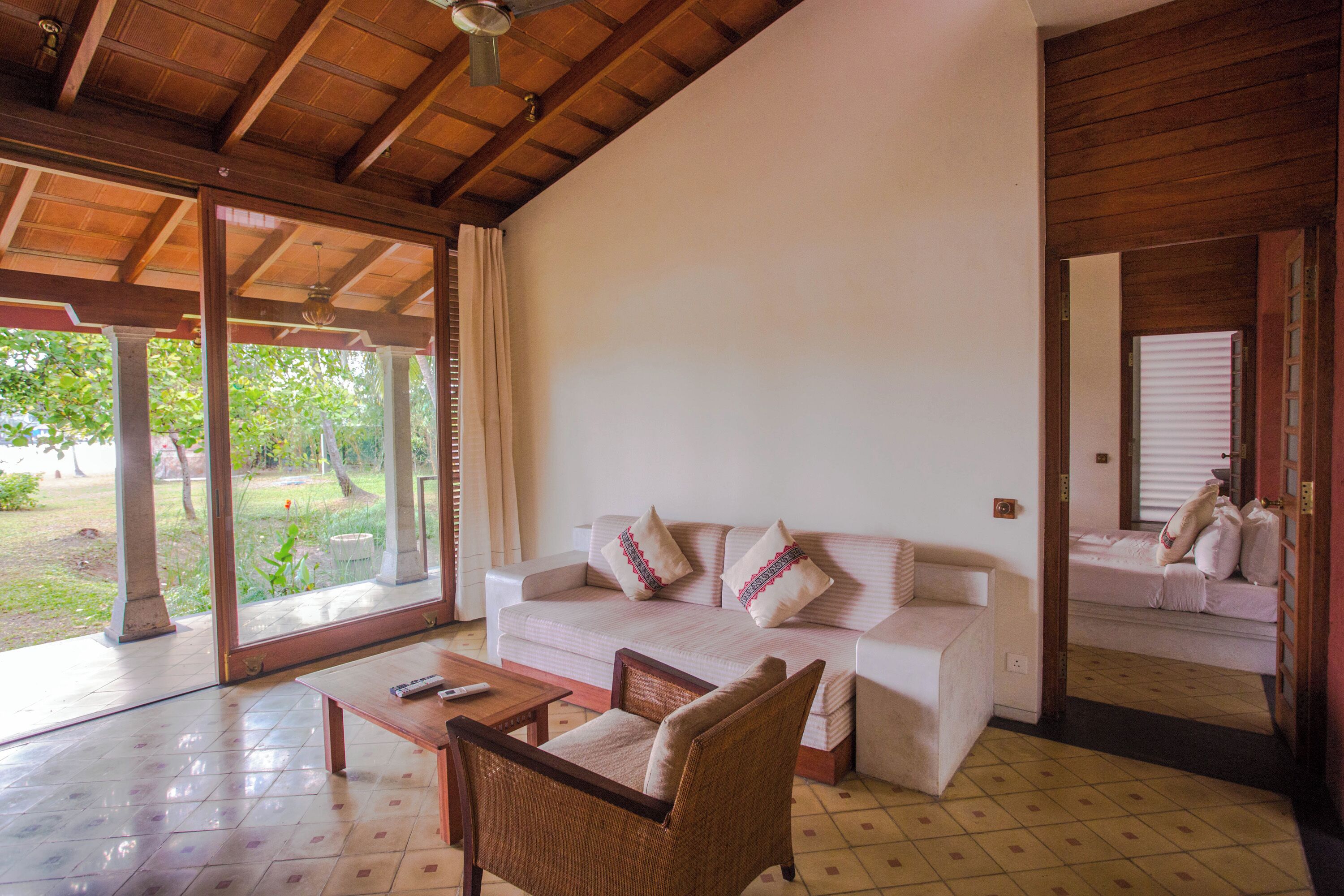 Amplia sala de estar con un sofá blanco, un sillón de mimbre y una mesa de centro. Grandes ventanales ofrecen vistas al exterior.