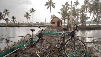 Una bicicleta estacionada cerca de una pequeña estructura de madera a orillas de un cuerpo de agua tranquilo, rodeado de palmeras y barcos pesqueros.