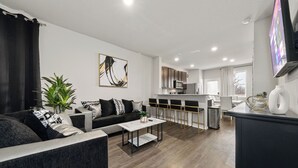 Una sala de estar moderna con un sofá, una mesa de centro y una planta. Hay una zona de cocina con armarios y un microondas.