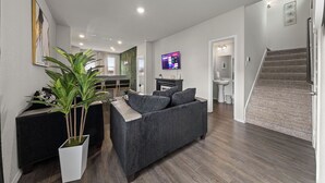 Una sala de estar moderna con un sofá gris, una maceta blanca con una planta, un televisor de pantalla plana y una escalera.