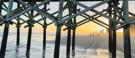 A pier structure with a view of the beach and distant cityscape at sunset.