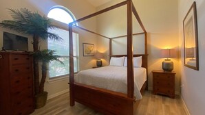 Un letto a baldacchino con struttura in legno, una televisione su un comò, una palma in vaso e una finestra con persiane.