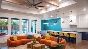 A modern living room with a large orange sectional sofa, green armchairs, and wooden coffee tables. A kitchen with blue cabinets and yellow bar stools is visible in the background.