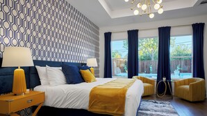 A modern bedroom with a patterned wall, a large bed with white and yellow bedding, two yellow armchairs, and a view of the outdoors.