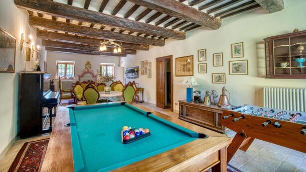Une table de billard avec des billes, une table de baby-foot, un piano et une table de snooker dans une pièce aux poutres apparentes et dont les murs sont ornés de tableaux encadrés.