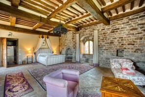 Une pièce avec un mur en pierre, un lit à baldaquin, un fauteuil violet, un tapis à motifs et une table basse en bois.