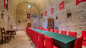 Une longue table entourée de chaises rouges, des murs en pierre et des drapeaux.
