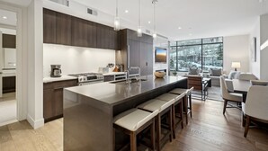 A modern kitchen with a large island, bar stools, and a dining area with a view of a snowy landscape. - Ski-In/Ski-Out Residence In The Heart Of Canyons Village (Park City)