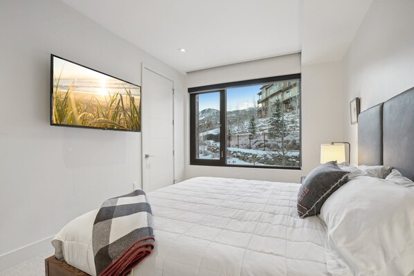 A bedroom with a large window, a bed, a bedside table, and a wall-mounted TV.