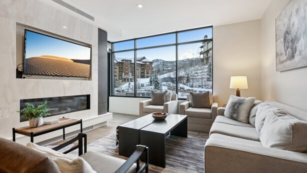 A modern living room with a large window, a flat-screen TV, a sofa, a coffee table, and a lamp. - Ski-In/Ski-Out Residence In The Heart Of Canyons Village (Park City)