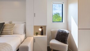 Un dormitorio con una cama, una silla, una mesita de noche con lámpara y una ventana con vistas a zonas verdes.