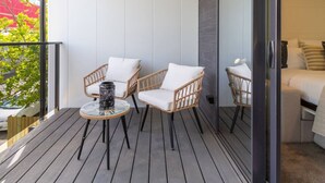 Un balcón con sillones de mimbre y una mesa de vidrio, una cama con ropa blanca y vistas a un edificio rojo.