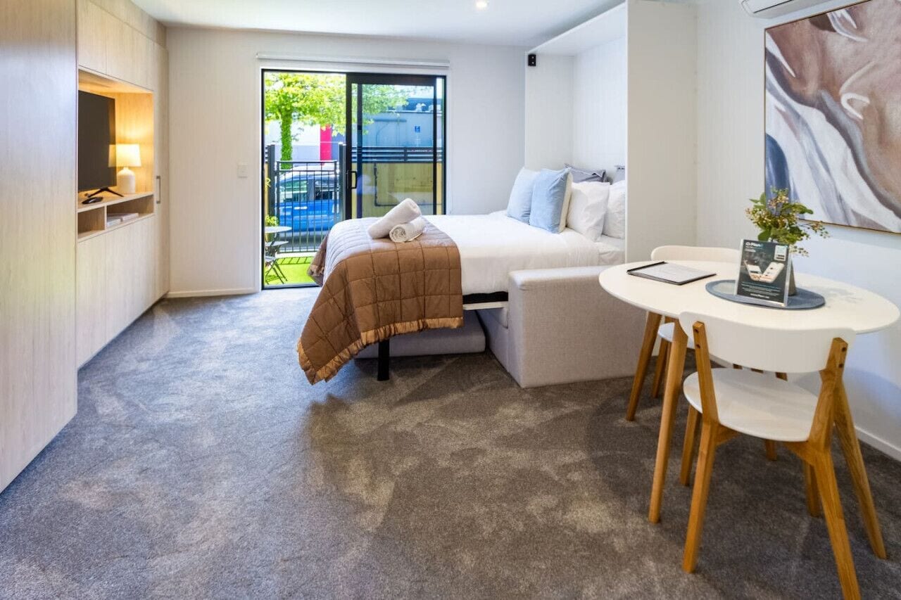 Ein modernes Hotelzimmer mit Bett, einem kleinen Tisch mit Stühlen, einem Fernseher und einer Glastür, die zu einem Außenbereich führt.