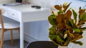 Ein moderner Arbeitsplatz mit einem weißen Schreibtisch, einem Computer, einer Topfpflanze und einer Vase mit grünen Blättern auf einem Tisch.