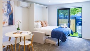 Una habitación de hotel moderna con una cama, una pequeña mesa con sillas, una planta en maceta y un ventanal con vistas al exterior.