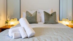 Ein ordentlich bezogenes Bett mit weißen Leinen und zwei Kissen, ein Nachttisch mit einer Lampe und einer Blumenvase sowie ein großer Spiegel an der Wand.