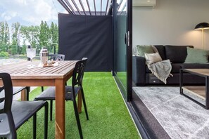 Un espace extérieur moderne aménagé avec une table et des chaises en bois, un canapé noir et une cloison en verre.