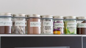 Una repisa con frascos etiquetados con diversos ingredientes como 'Plunger Coffee', 'Té Negro', 'Chocolate Bebible', 'Azúcar', 'Douve Egberts', 'Té Dulce', 'Menta' y 'Café'.