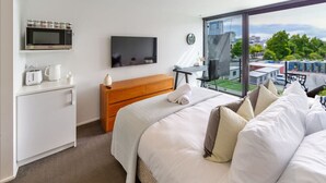 Una habitación de hotel moderna con una cama grande, una mesita de noche de madera, un televisor de pantalla plana, una cafetera y vistas a la ciudad a través de amplios ventanales.