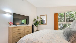 Ein Schlafzimmer mit einem Bett, einem Fernseher auf einer Holzkommode, einem Spiegel und einem Fenster mit Blick auf Bäume.