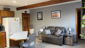 A compact living space with a kitchenette, a gray sofa with cushions, a dining table with two chairs, and a small side table with a lamp.