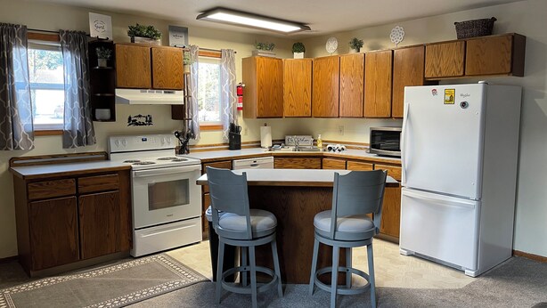 A compact kitchen with wooden cabinets, a white refrigerator, a stove, and two bar stools.