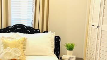 A neatly made bed with pillows, a bedside table with a plant and a book, and a window with curtains.