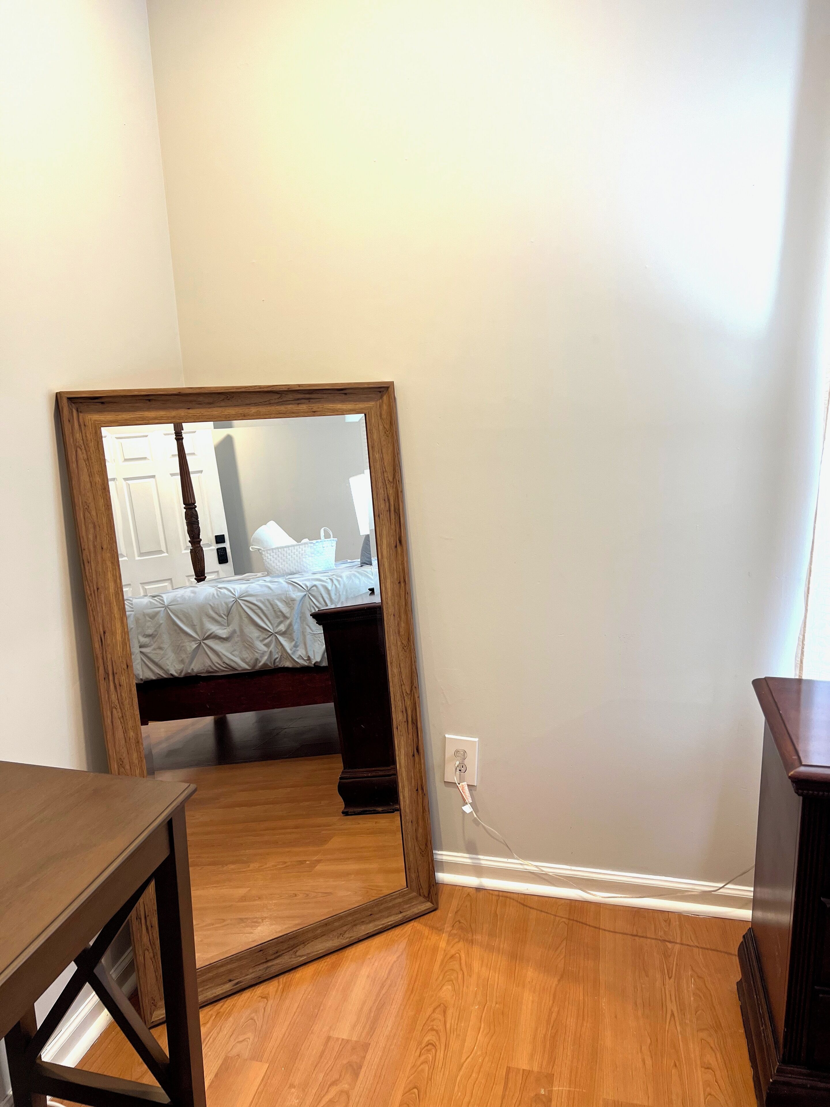 A bedroom with a wooden floor, a bed with white bedding, a nightstand, and a mirror leaning against the wall.